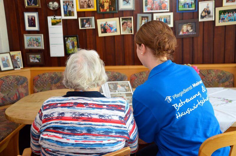 Eine Pflegefachkraft schaut zusammen mit einer Seniorin ein Fotoalbum durch. Sie sind von hinten fotografiert