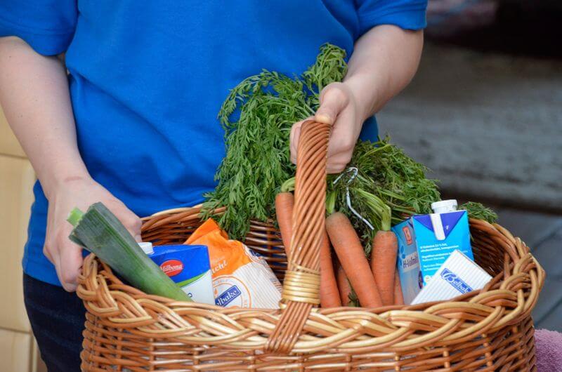 Eine Mitarbeiterin hält einen Einkaufskorb mit Lebensmitteln in ihm: Milch, Möhren, Zwieback, Porree