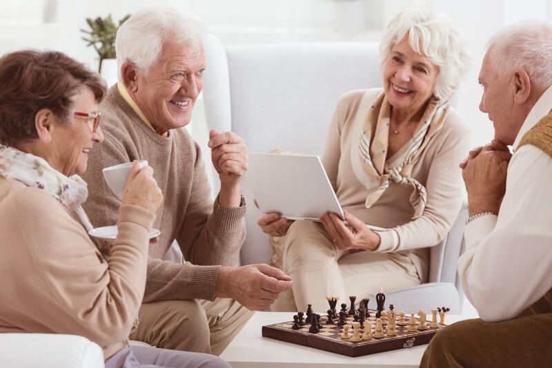 Zwei Senioren und zwei Seniorinnen sitzen im Wohnzimmer gemeinsam an einem kleinen Couchtisch und trinken Kaffee, spielen Schach und lachen.