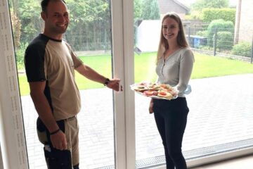 Das Foto zeigt einen Handwerker an der Terrassentür. Neben ihm steht eine junge Frau mit einem Tablett mit belegten Brötchen in der Hand.