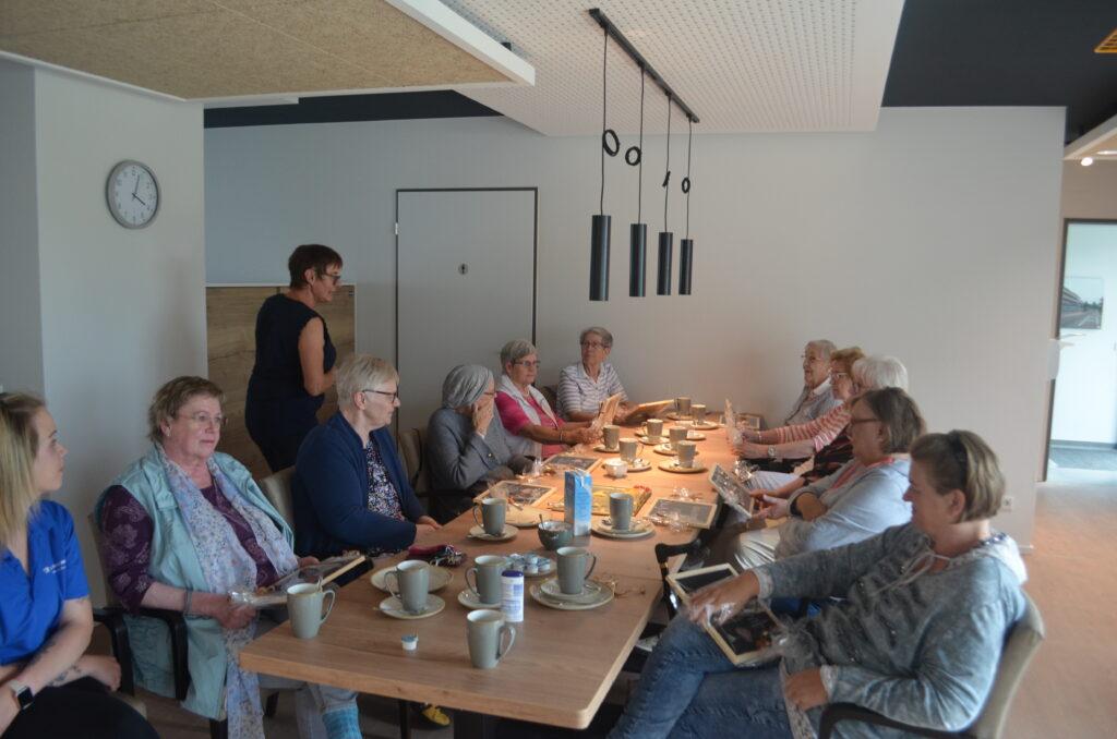 10 Seniorinnen und zwei Pflegefachkräfte sitzen an einem langen Tisch,trinken Kaffee und schreiben etwas auf kleine Tafeln.